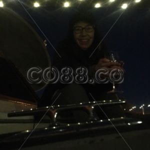 Older Woman enjoying a Nightcap while sitting on her Vessel’s Deck with Lights reflecting on Cleat - CO88.co